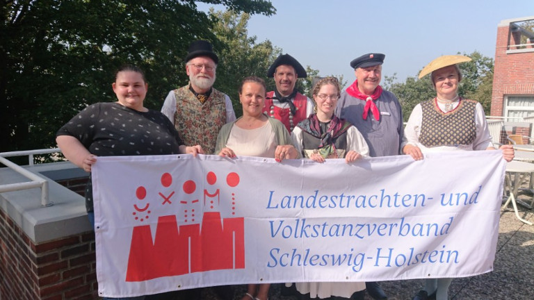Sieben Personen mit Banner "Landestrachten- und Volkstanzverband Schleswig-Holstein"