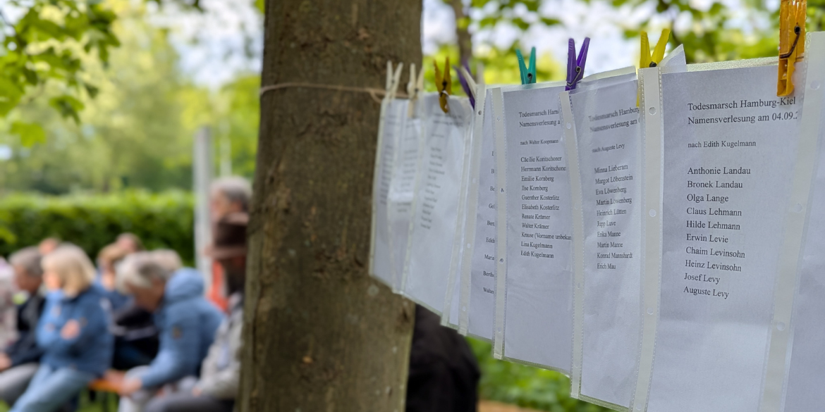 Zettel mit Namen von Teilnehmenden des Todesmarsches an Wäscheleine, im Hintergrund Personen