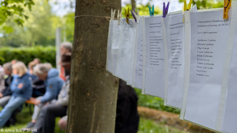 Zettel mit Namen von Teilnehmenden des Todesmarsches an Wäscheleine, im Hintergrund Personen