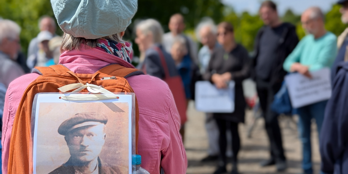 Frau mit Rucksack, auf dem ein historisches Porträtfoto eines Mannes klebt, im Hintergrund weitere Personen