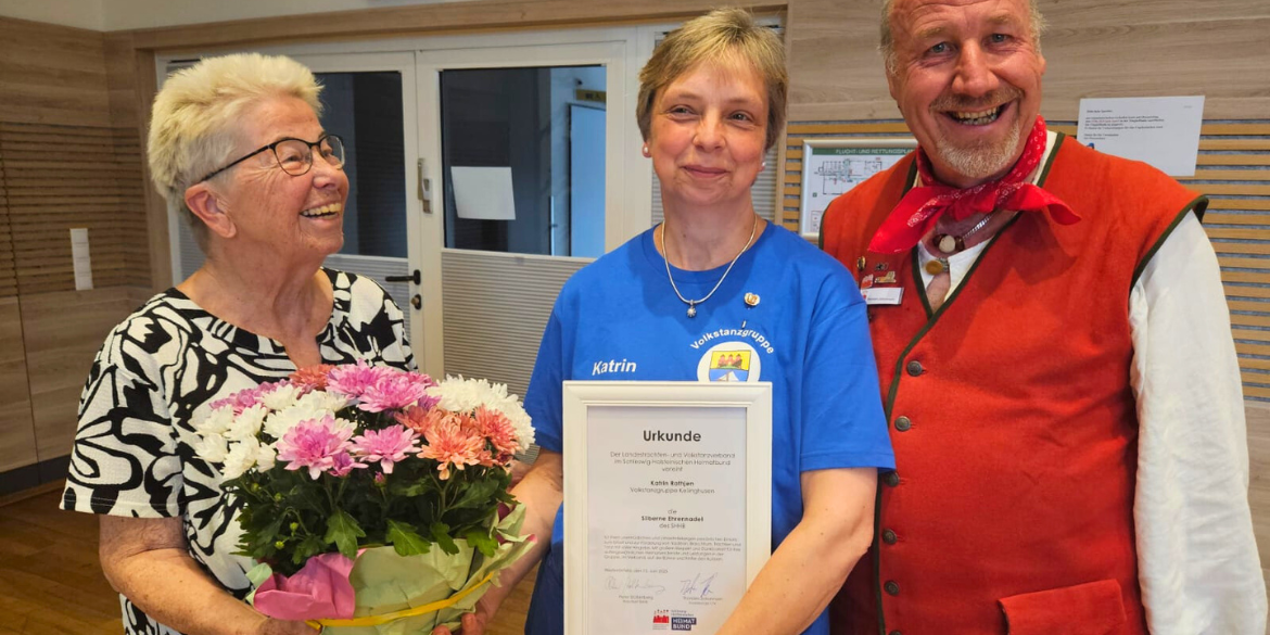 Drei Personen, eine Frau mit Blumenstrauß, eine mit Urkunde, ein Mann trägt Tracht
