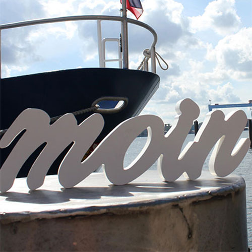Schild mit Aufschrift "moin" vor einem Schiff am Hafen